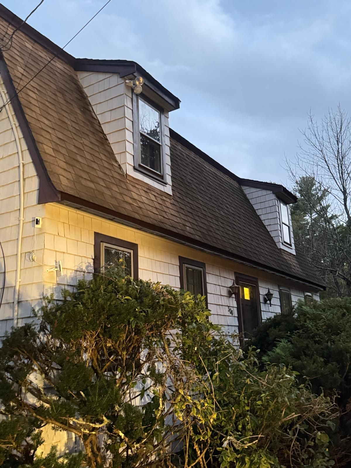 White house with brown roof and dormer windows, surrounded by green bushes.