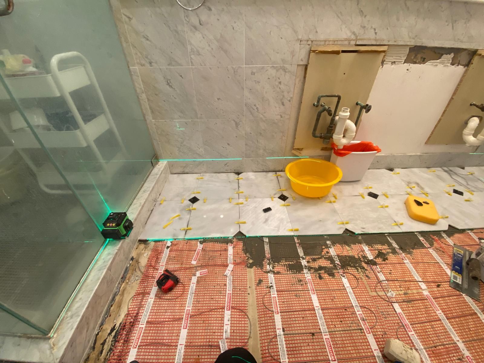 Bathroom floor renovation with laser level projecting green lines.  Heated floor mat, tiles, and tools are visible.