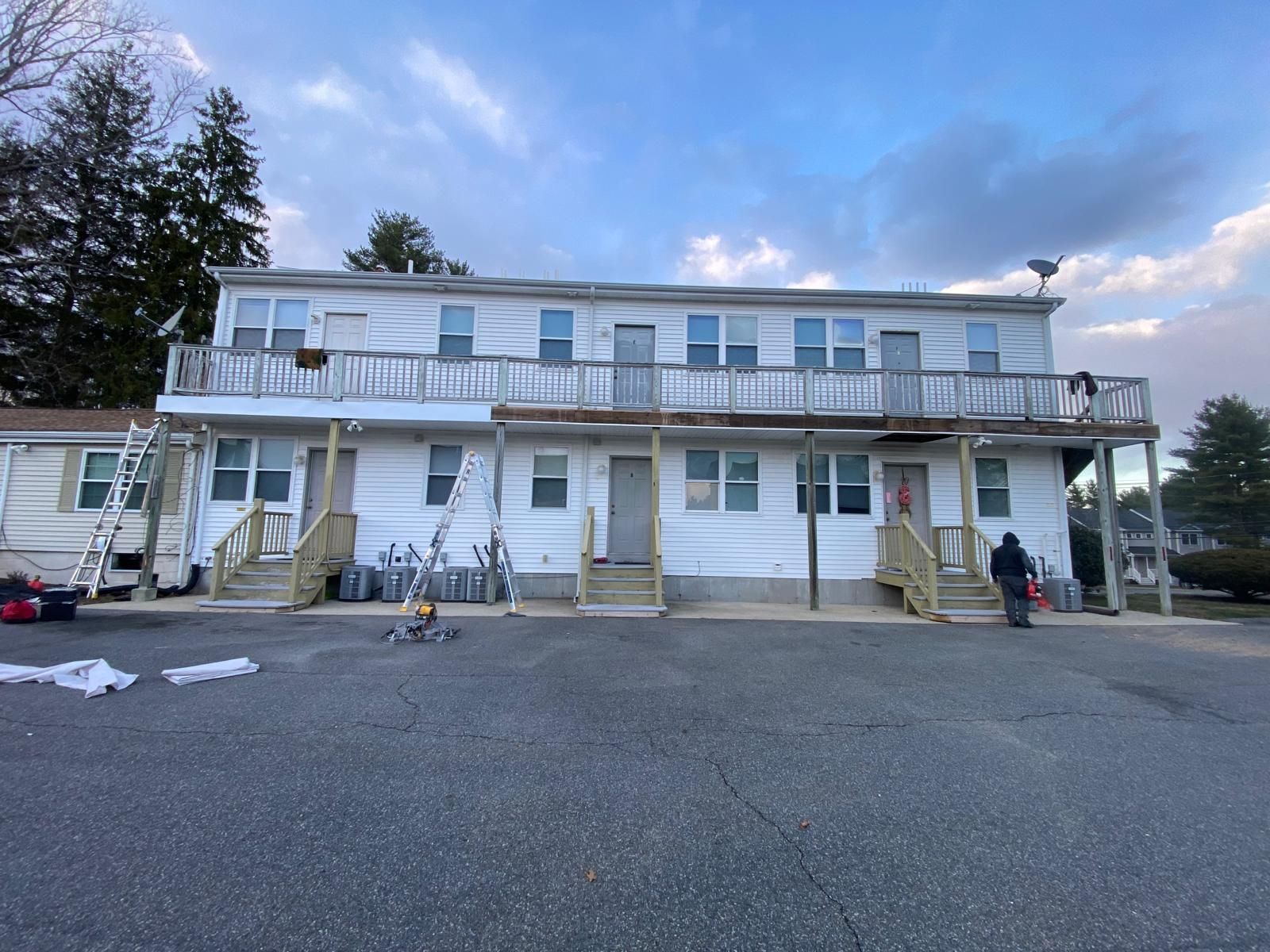 Two-story white building with wooden balconies; construction underway. Paved area in front; cloudy sky.