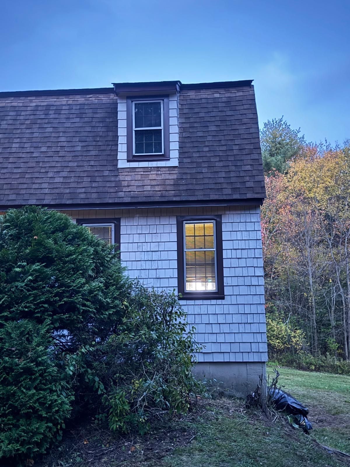 Two-story house with weathered shingles, small window in a dormer, two windows lit, set among trees and bushes.