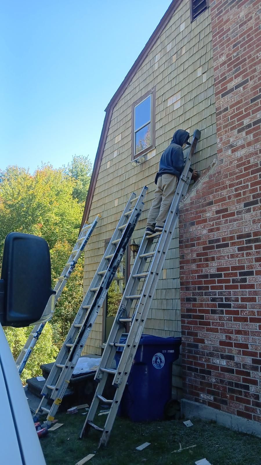 A person on a ladder repairs siding on a house next to a brick chimney, blue sky overhead.