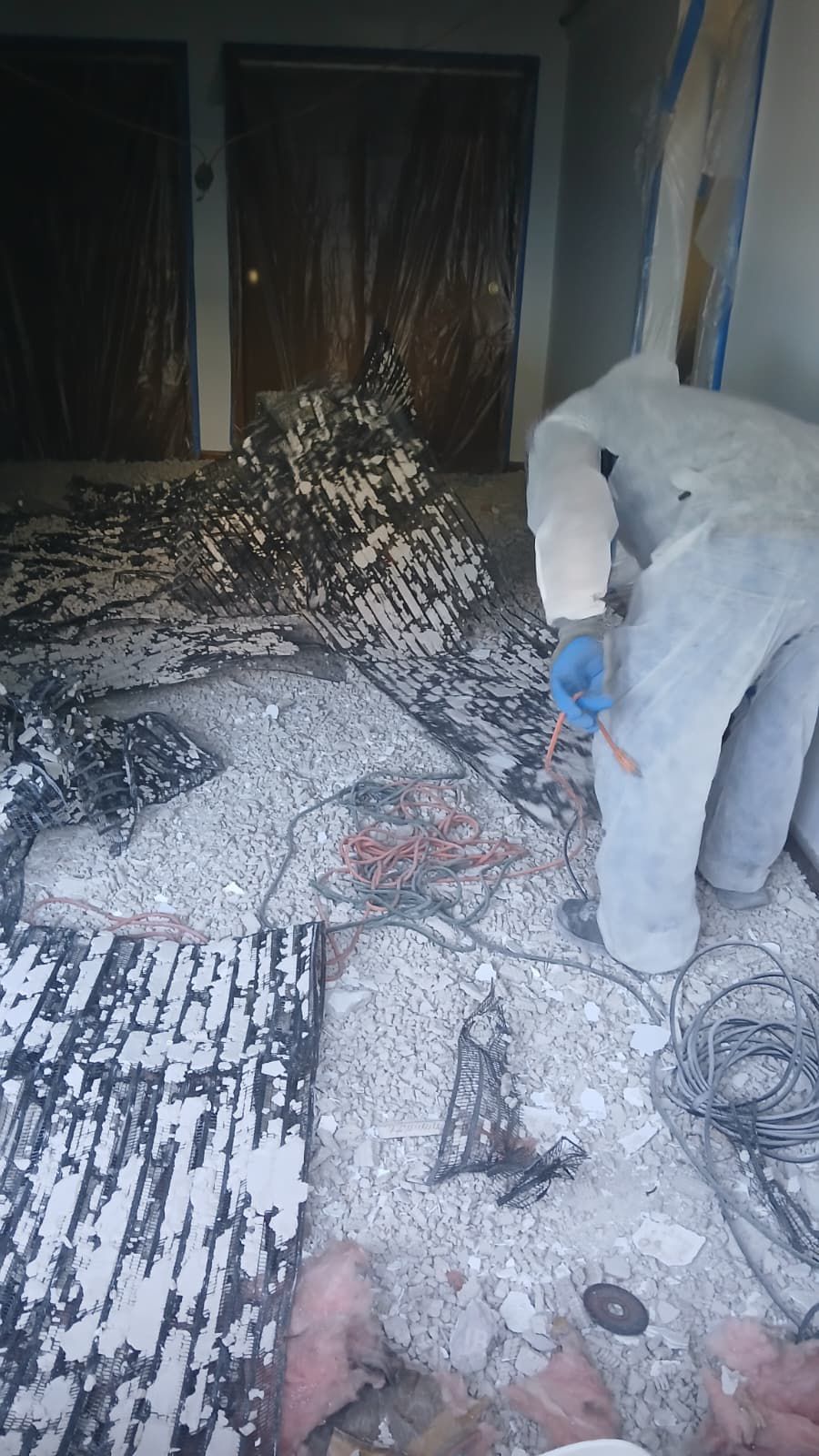 Construction site. Person in protective suit inspects debris-covered room with two doors in the background.