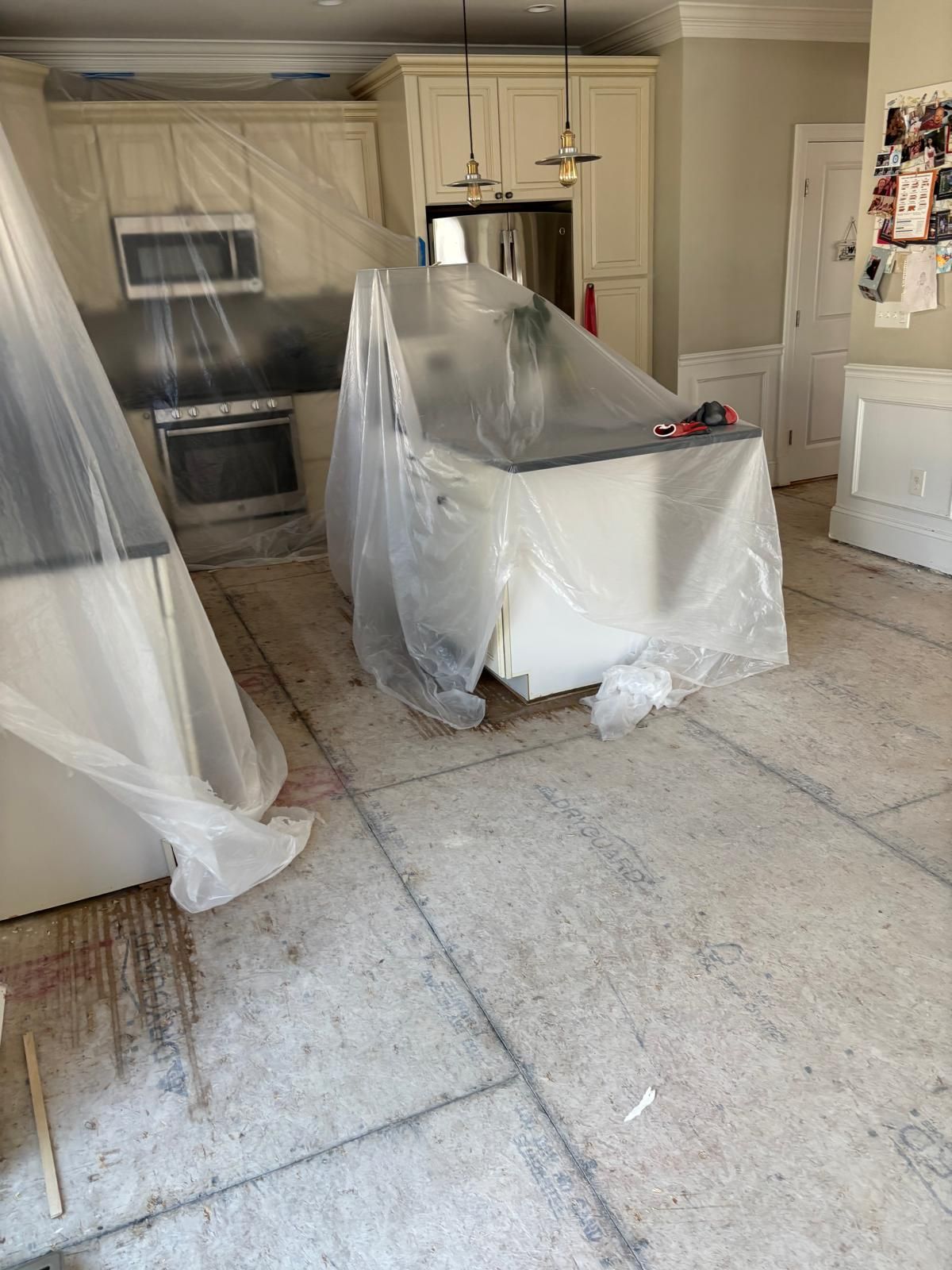 Kitchen renovation: Cabinets, appliances, and island covered in plastic sheeting. Old flooring exposed.