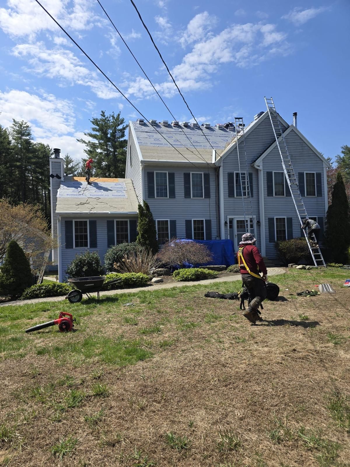 Roofers on a house roof. A blue house has a partly torn roof. A ladder and equipment are on the lawn.