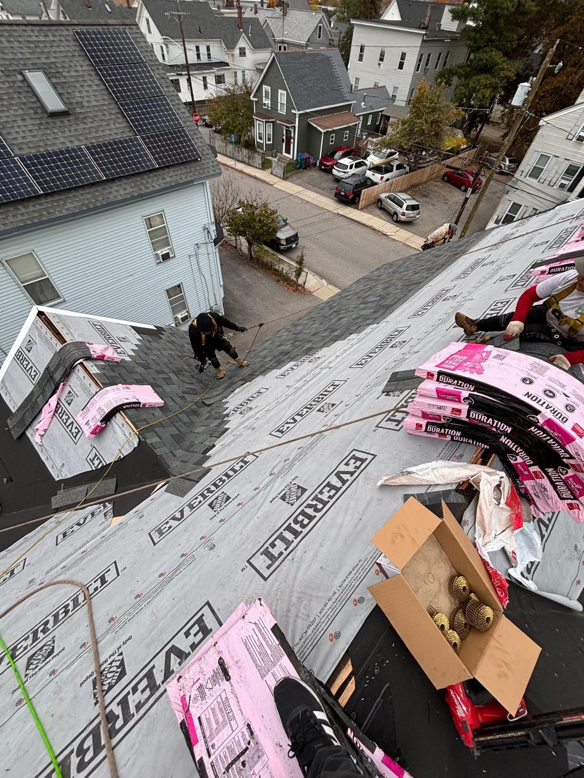 Roofers working on a residential roof, asphalt shingles and underlayment visible. City street and buildings in background.