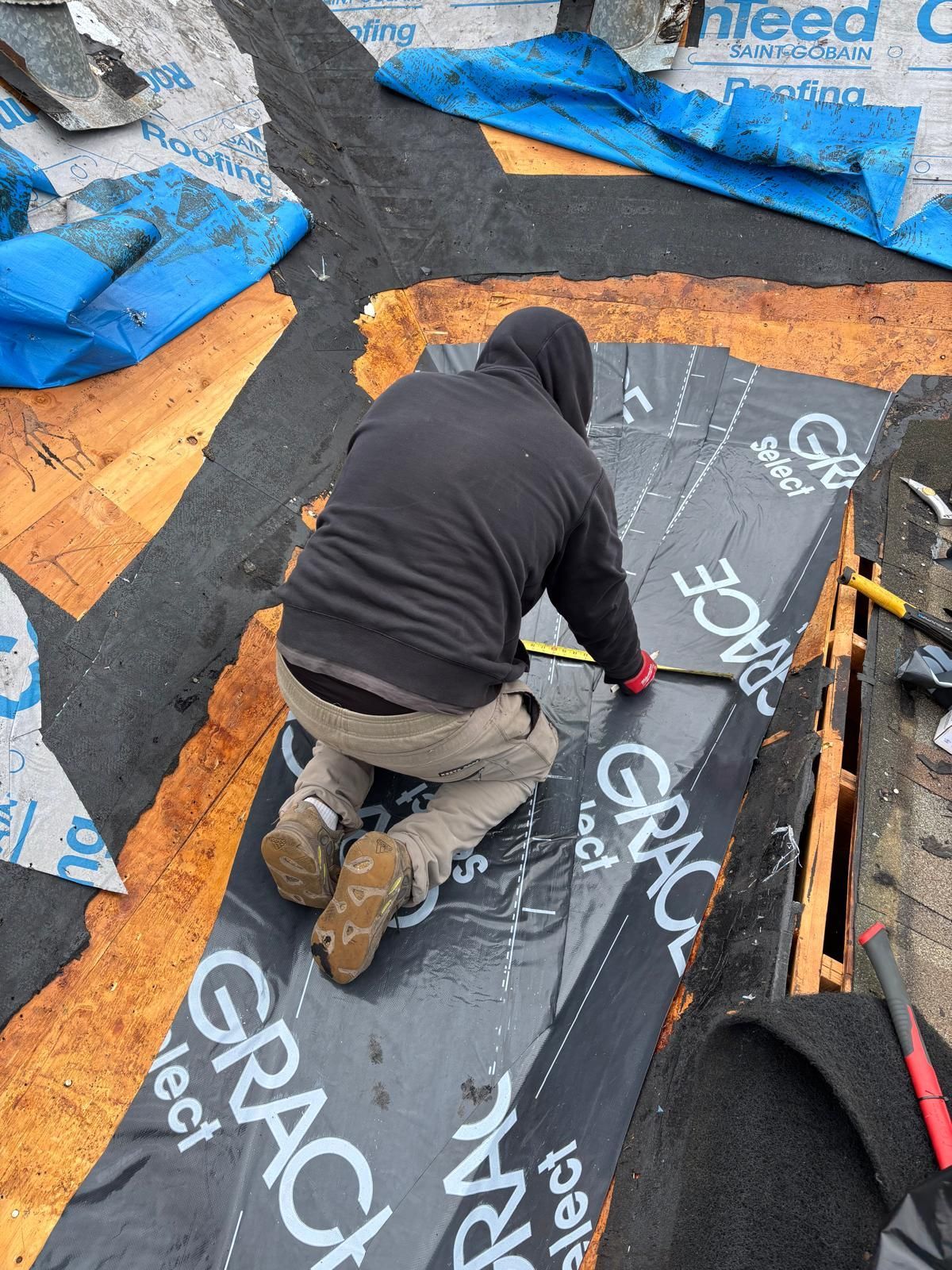 Person on a roof measuring a black membrane. Black asphalt roof with blue tarp visible.