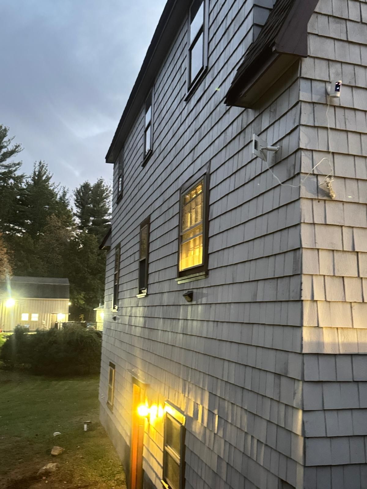 Side view of a two-story building with gray shingle siding, illuminated windows, and a grassy yard.