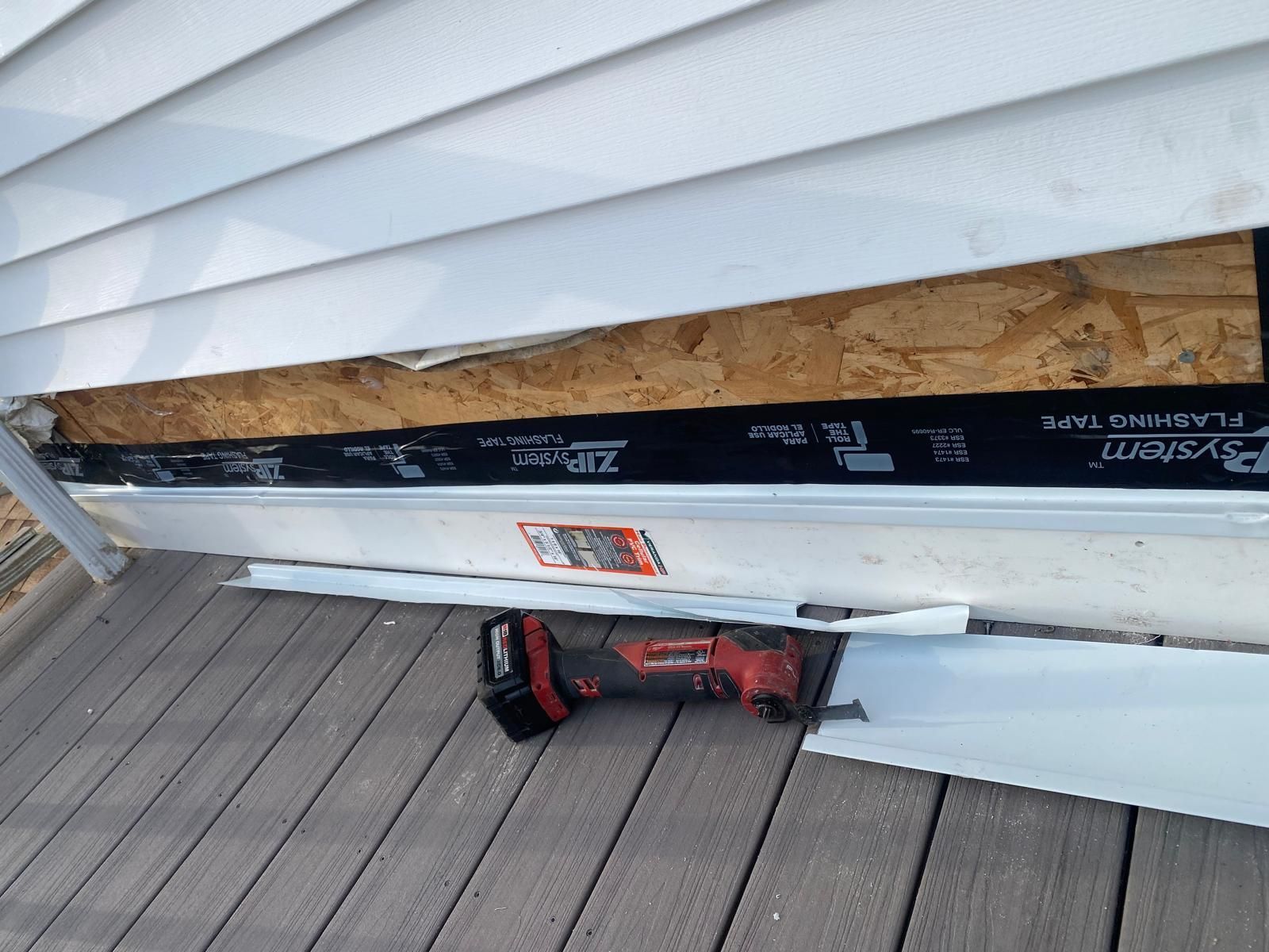 White siding being repaired, exposing wood and black flashing tape. A power tool rests on a wooden deck.