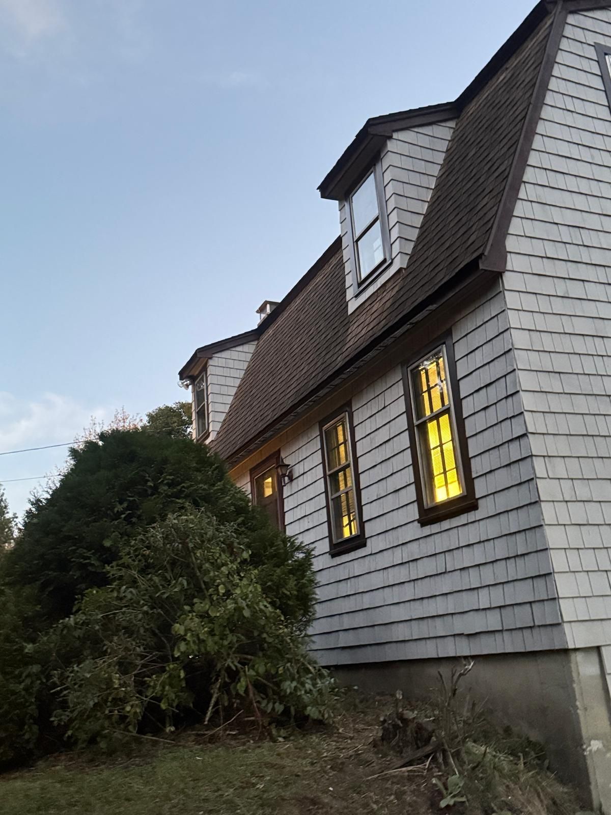 Side view of a gray shingle house with a dark roof and dormer windows, lit from within.