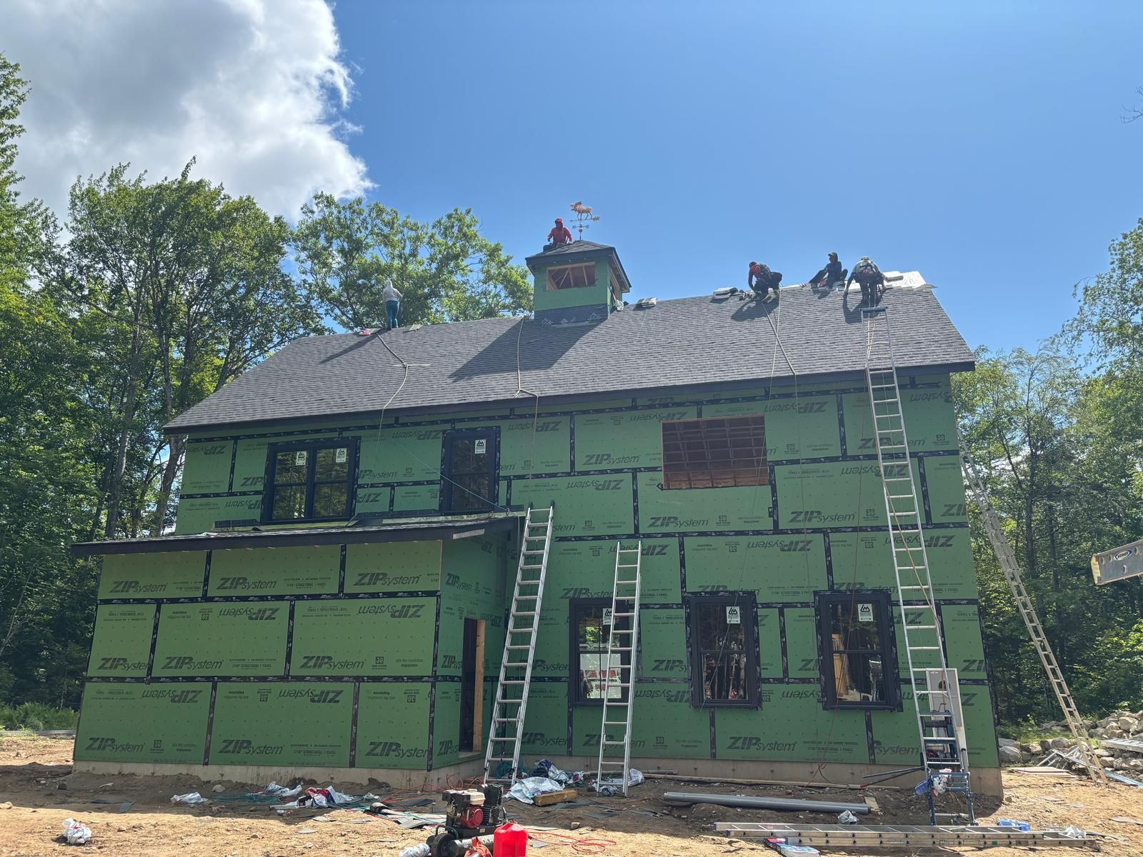 Construction workers on a roof, installing shingles on a two-story house with green siding, sunny day.