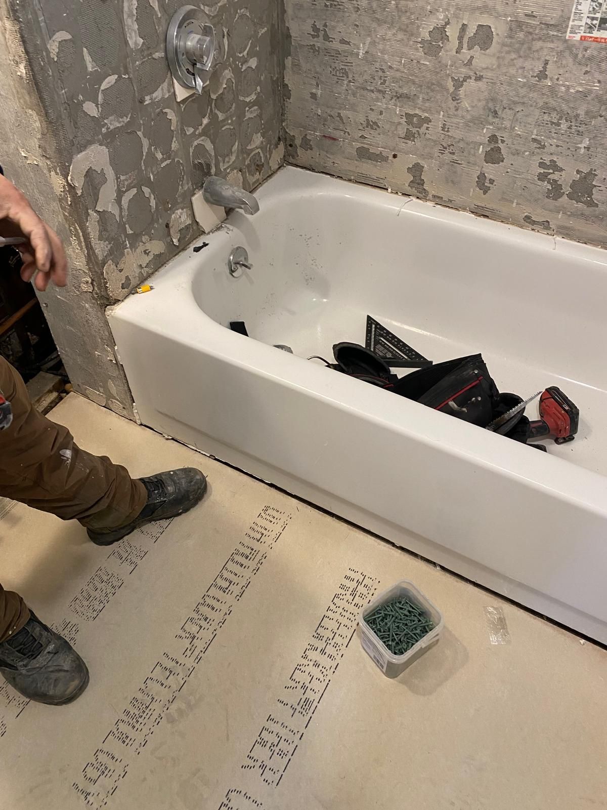 A white bathtub installed against a concrete wall. A person stands near it. Flooring is partially installed.