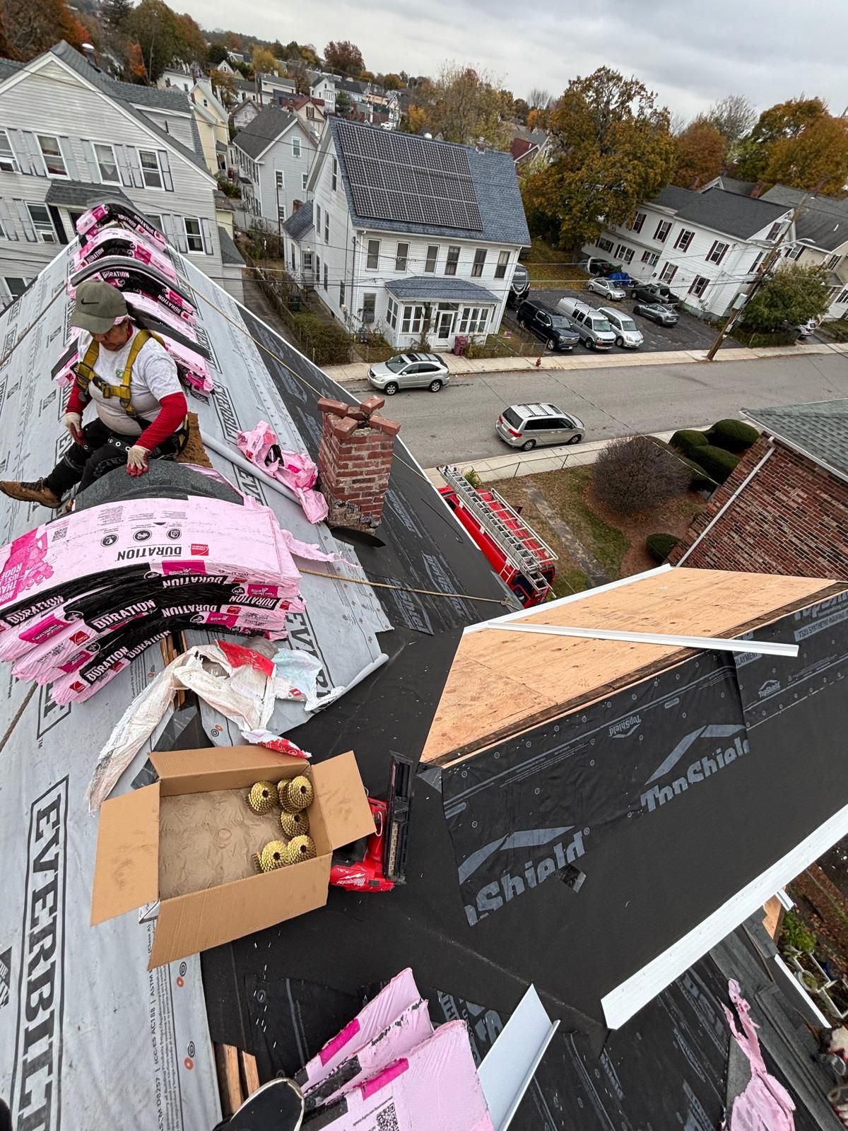 Roofer working on a roof, surrounded by shingles and materials. Buildings and cars visible in the background. Cloudy day.