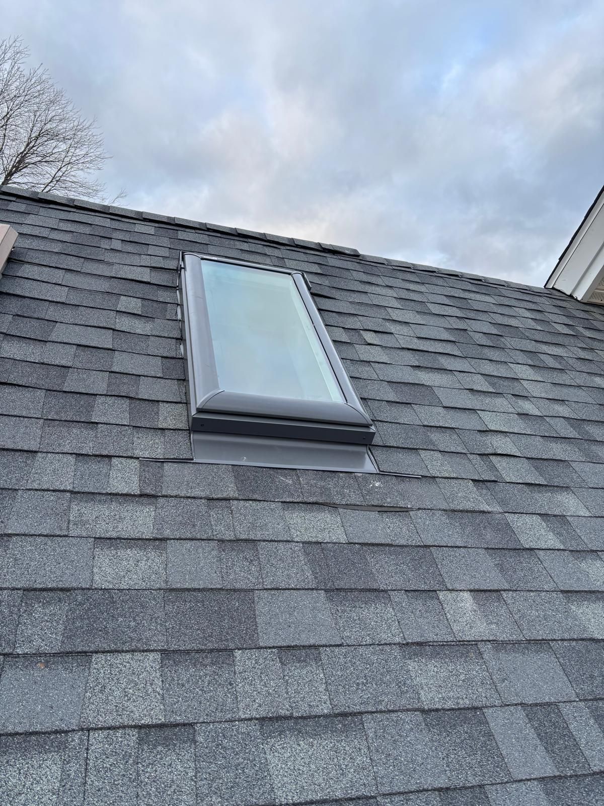 Skylight on a gray asphalt shingle roof against a cloudy sky.