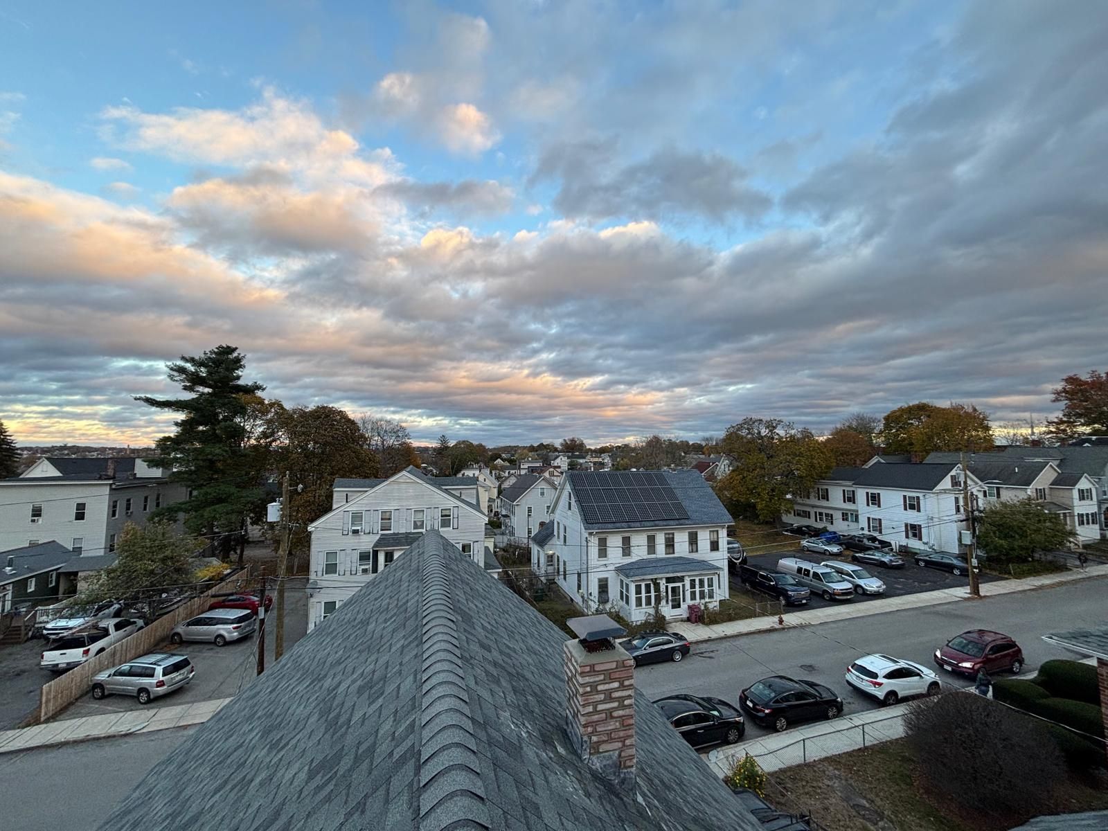 Rooftop view of a neighborhood street with houses, parked cars, and a cloudy sky at sunset.