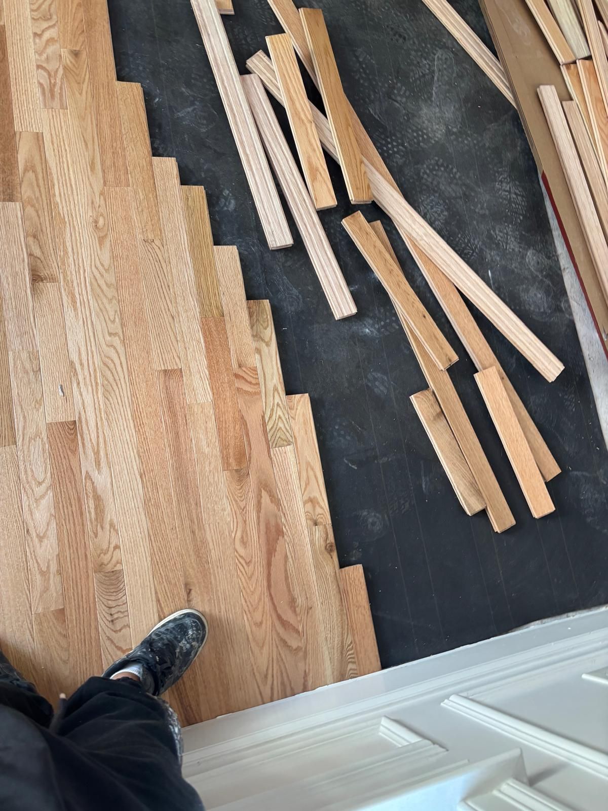 Hardwood flooring installation: rows of installed planks next to scattered pieces, person's foot visible.