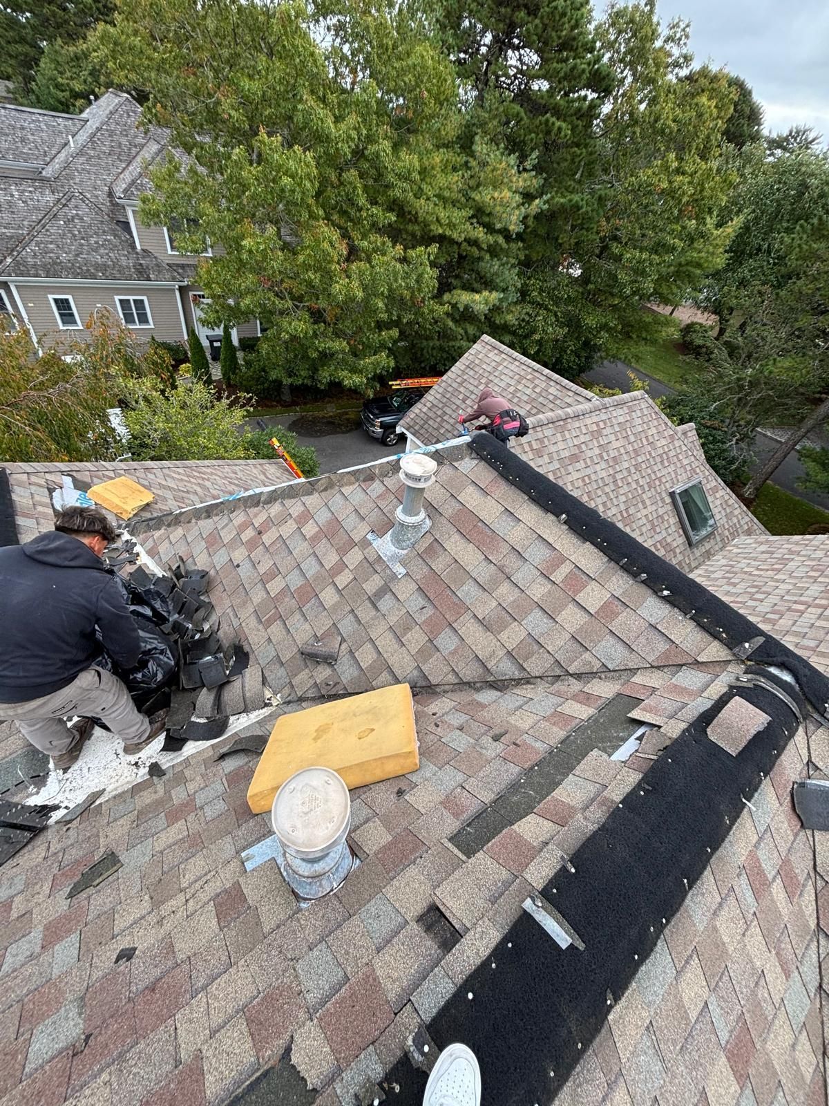 Roofers repairing a shingled roof with trees in the background. Tools and materials are scattered around.