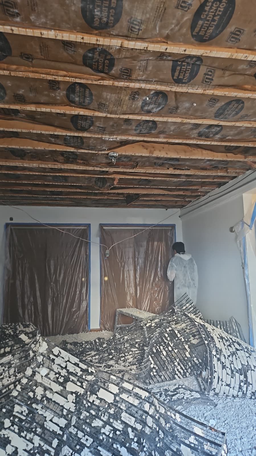 Room with exposed ceiling beams and debris on the floor; person in protective suit stands in doorway.