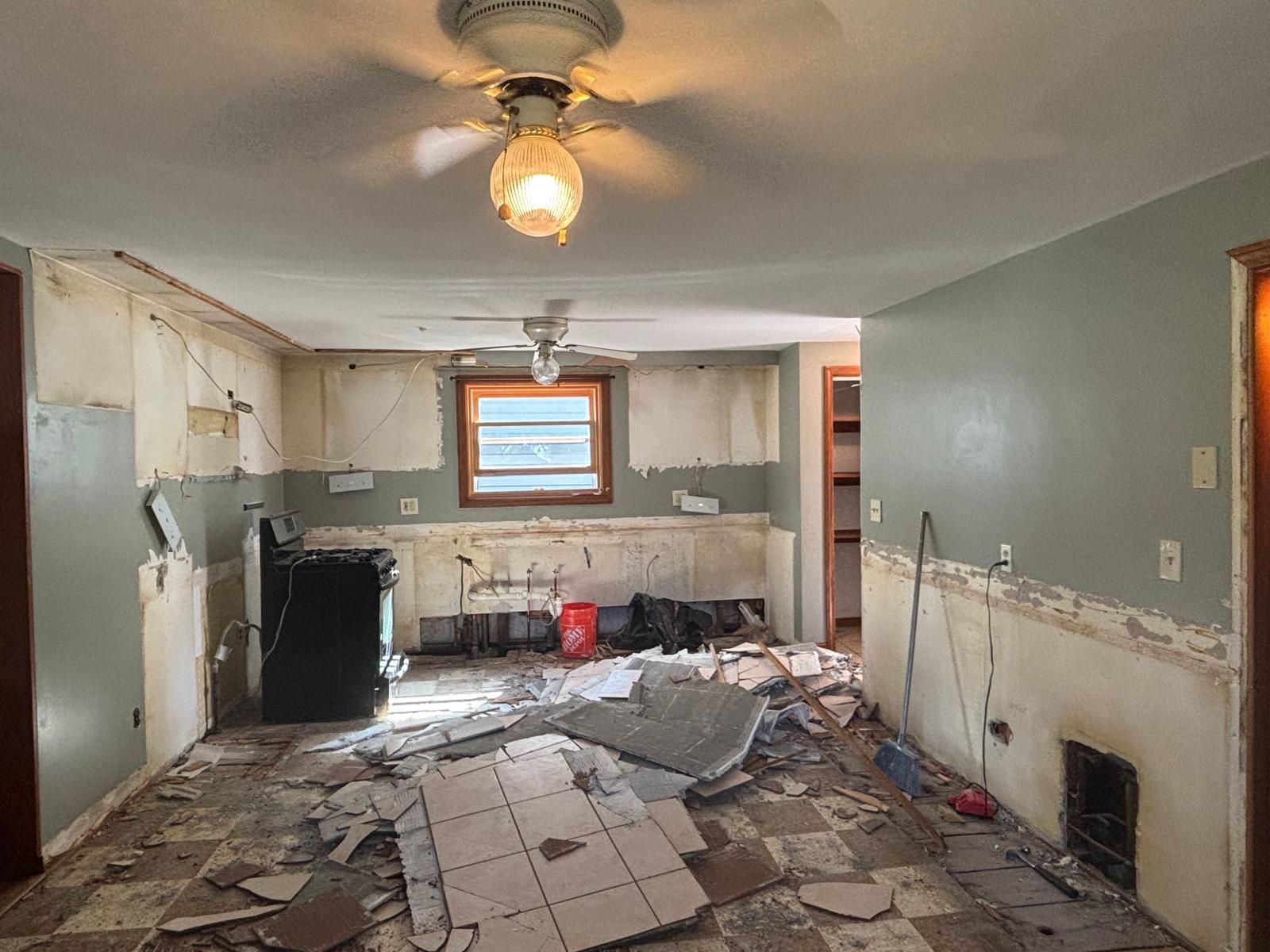 Kitchen renovation in progress; walls torn, flooring debris, cabinets removed, black stove, ceiling fans.