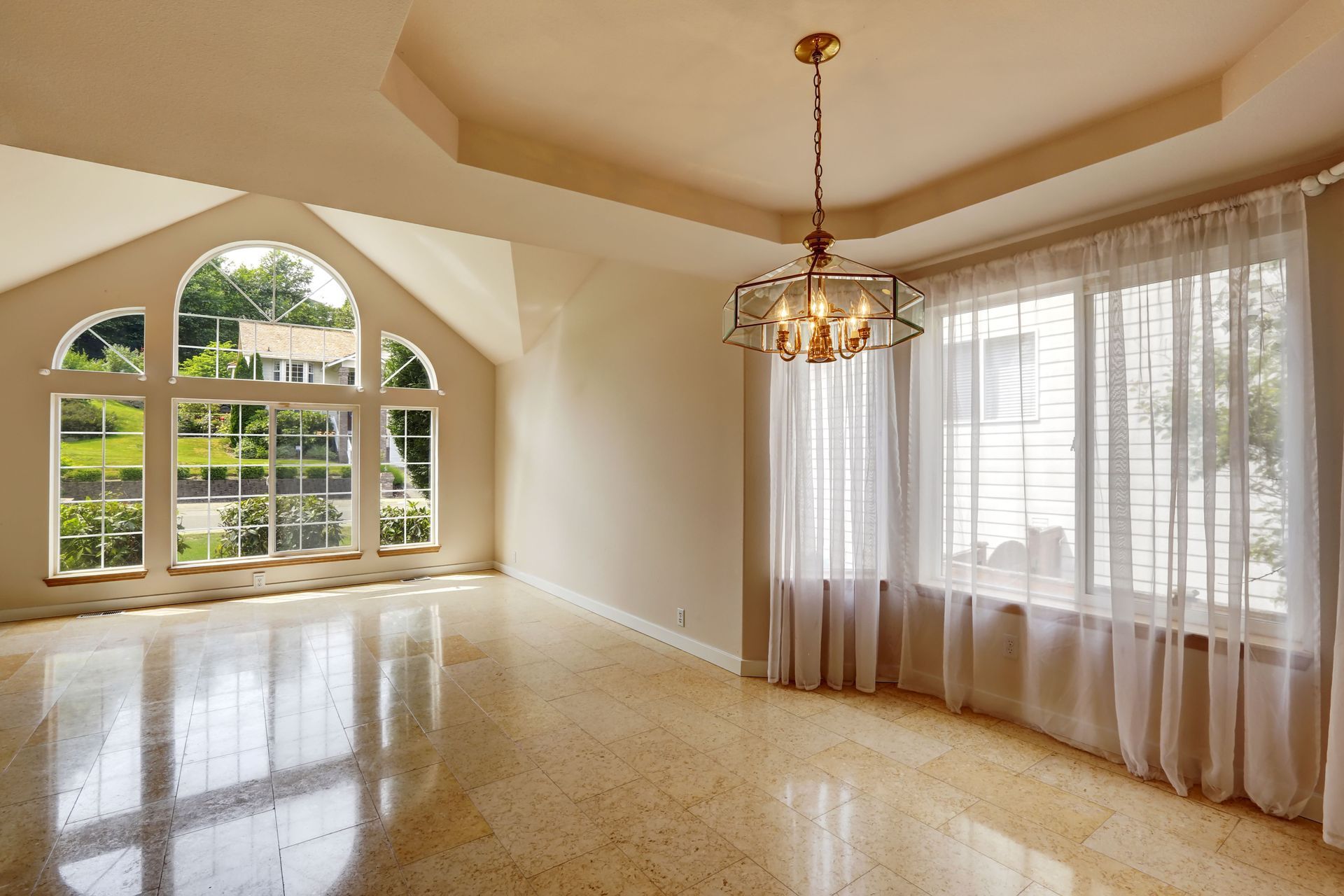 Empty, sunlit room with large windows and chandelier. Polished floor and sheer curtains.