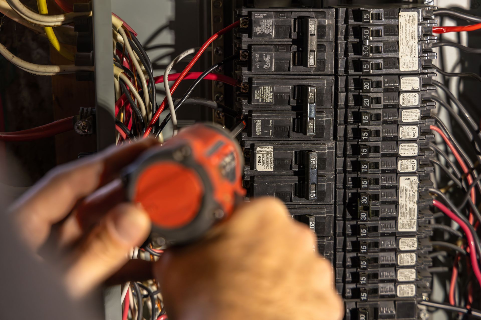 Electrician working on a circuit breaker panel with an orange screwdriver, wires visible.