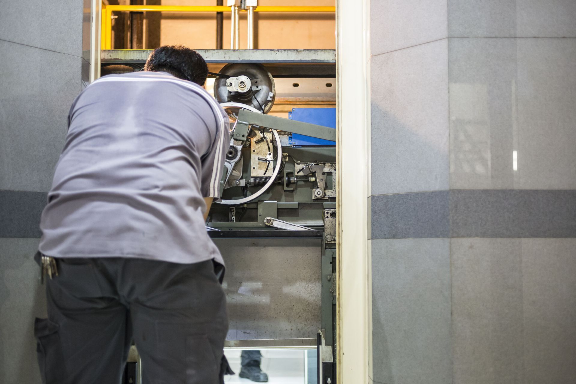 Man repairing elevator machinery. Gray shirt, dark pants, working inside the elevator shaft.