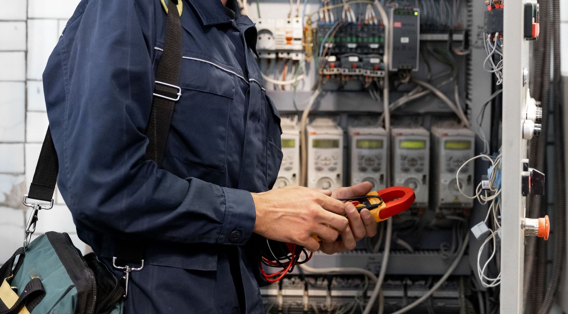 Electrician using a clamp meter on electrical panel. Blue jumpsuit, toolbox, wires visible.