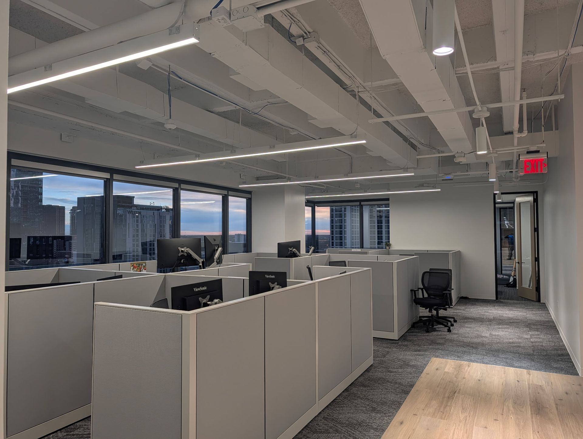 Office cubicles with computers and chairs, city views through windows, overhead lighting, and an exit sign.