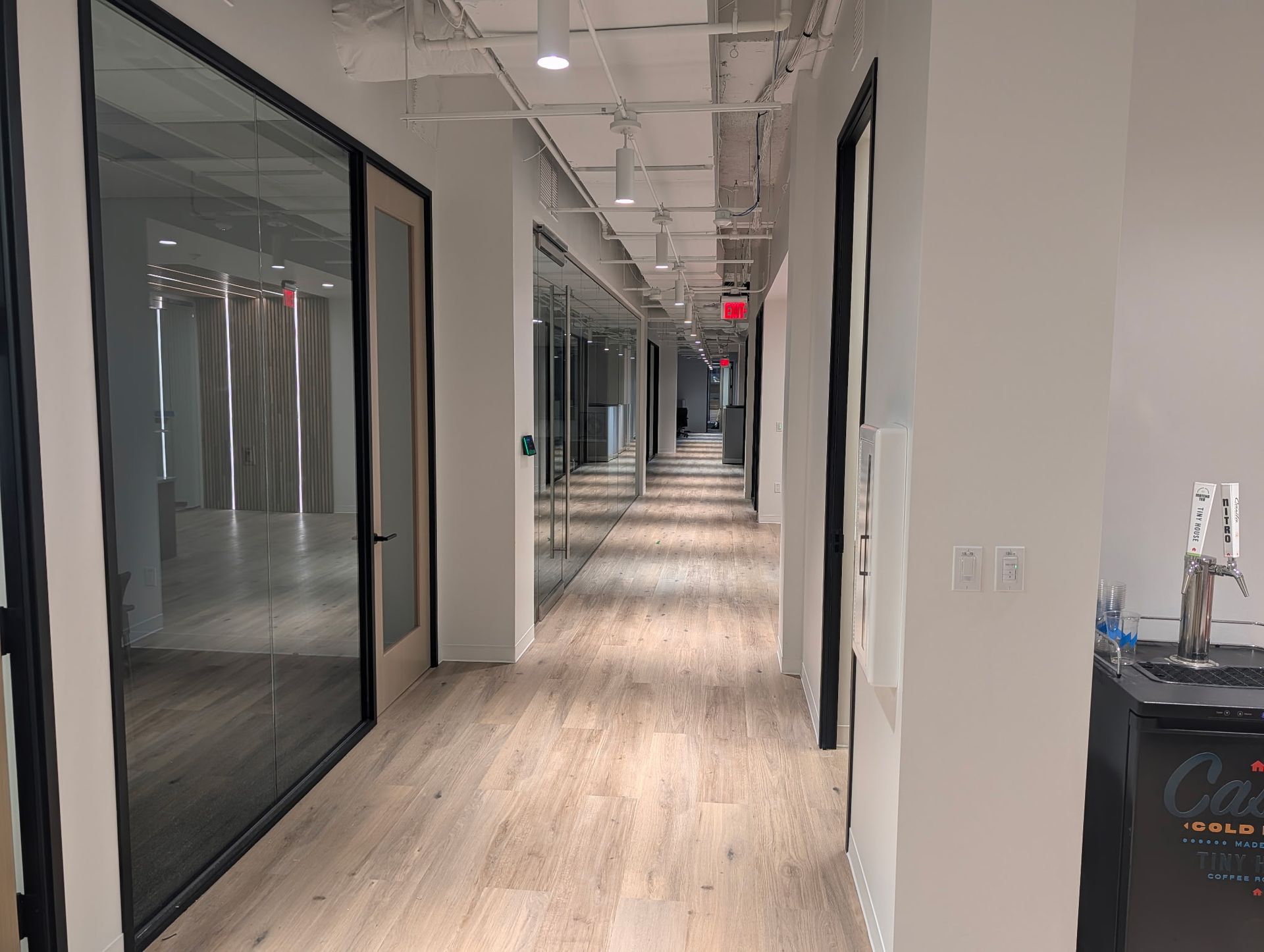 Office hallway with glass-walled rooms and light wood flooring, with a tap system on the right.