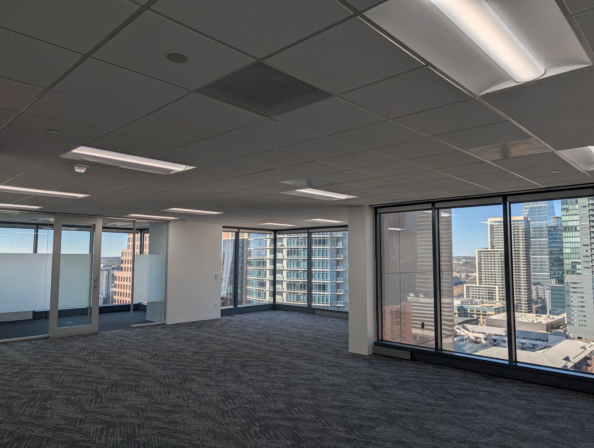 Empty office space with large windows overlooking a city skyline, gray carpet, and white walls.