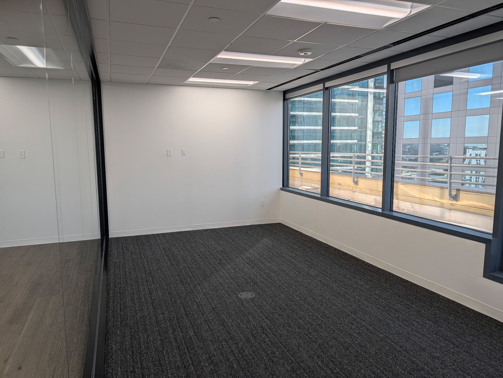 Empty office room with gray carpet, white walls, and large windows overlooking city buildings.