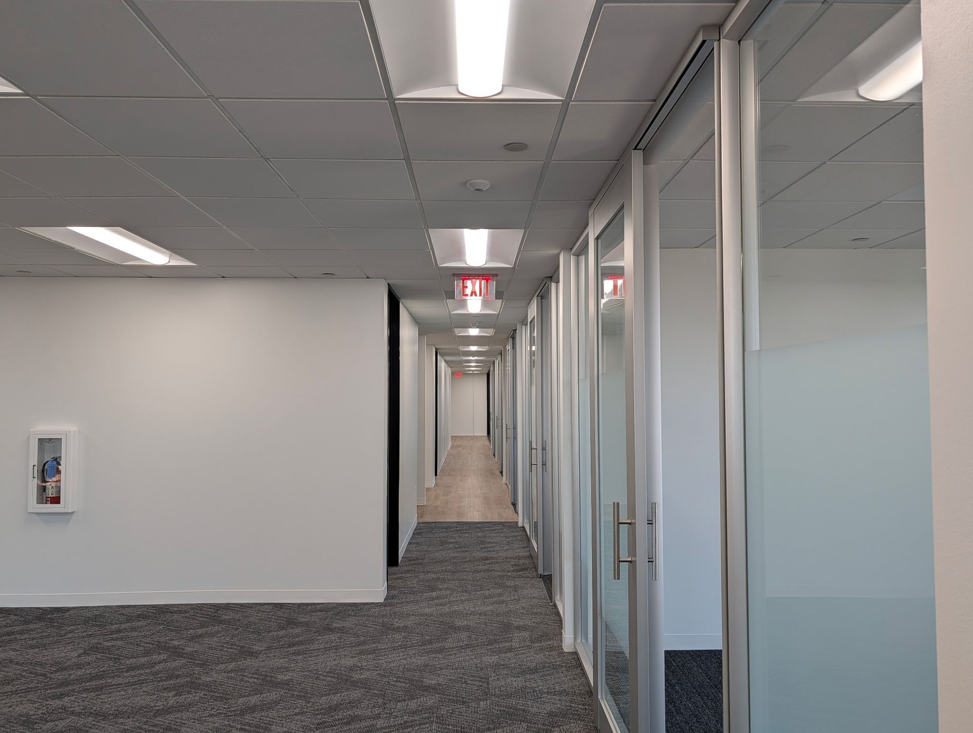 Empty office hallway with glass-walled offices on the right, exit sign visible down the hall.