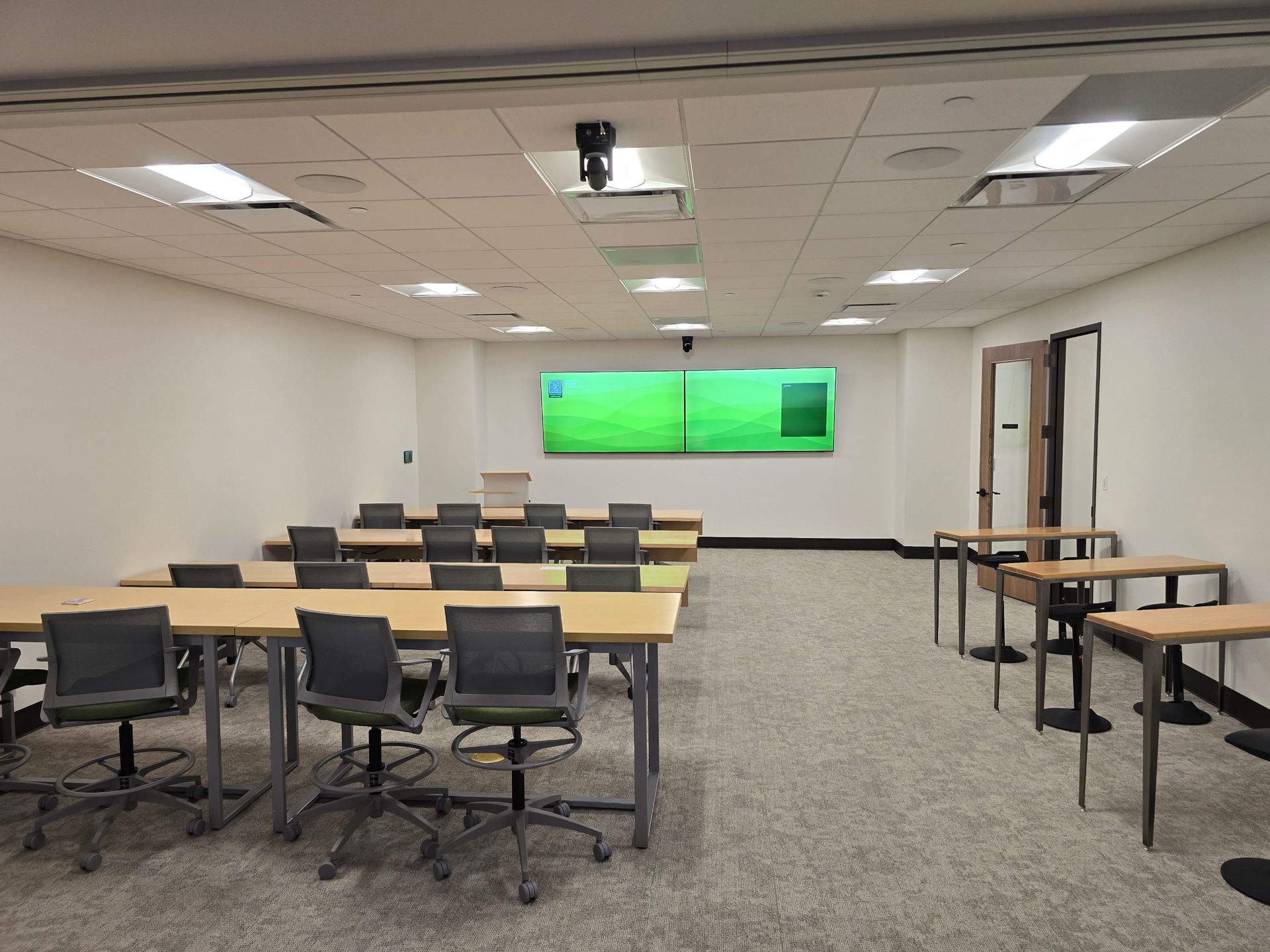 Classroom with rows of desks, chairs, and a large screen. White walls, drop ceiling, and gray carpet.