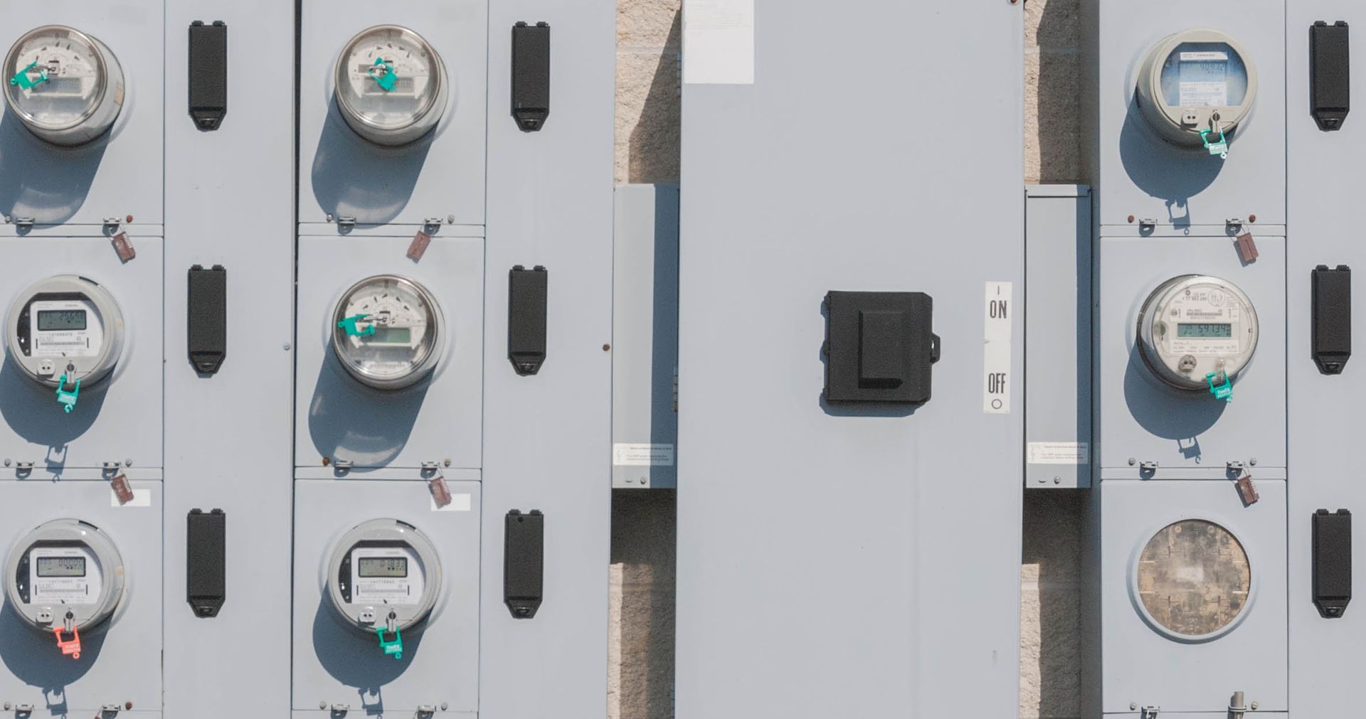 Gray electric meter boxes on a wall, each with a digital meter and black access panels.