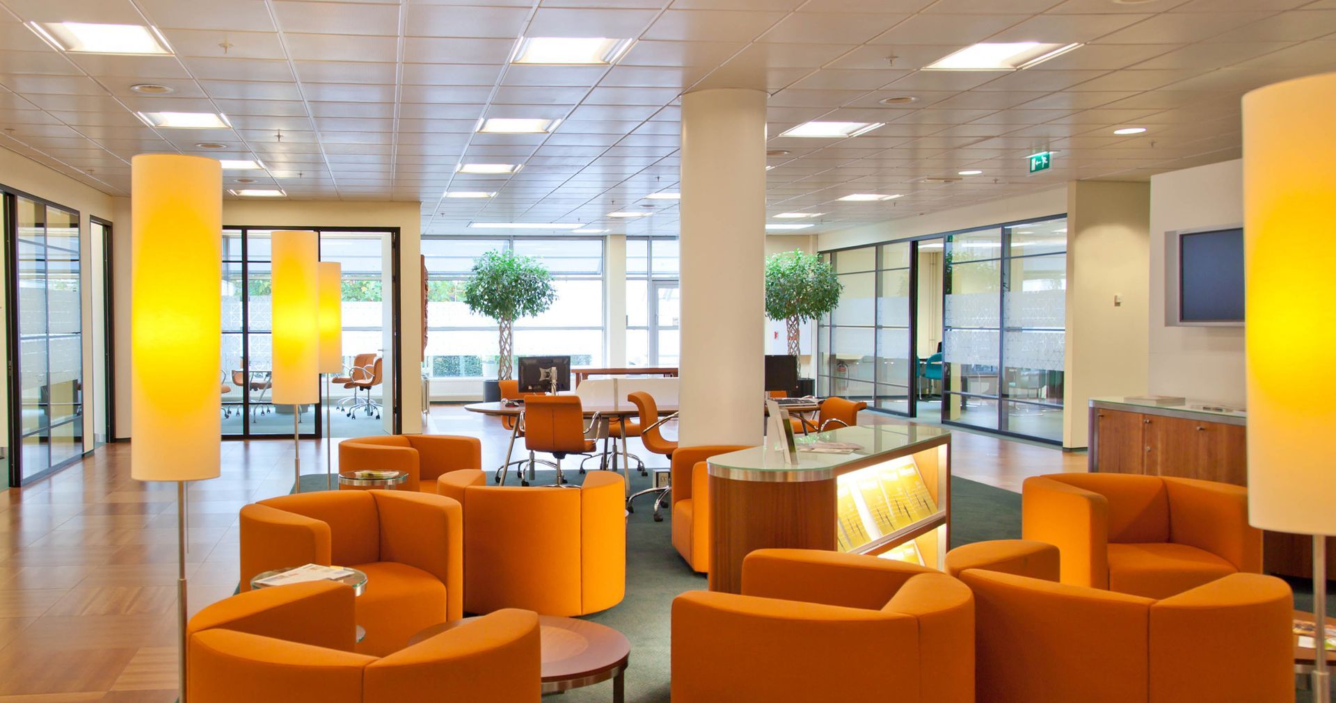 Orange lounge seating in a bright office space with floor lamps, glass-walled offices, and a central pillar.