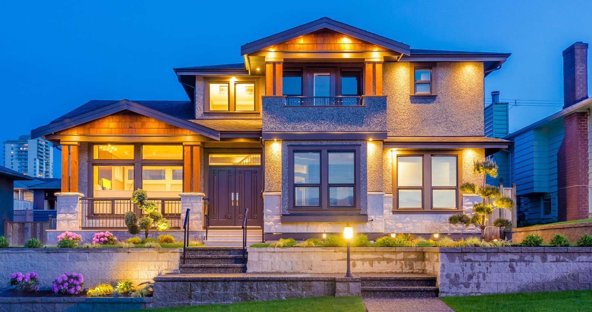Well-lit two-story house with dark roof, stone and wood accents, at dusk. Landscaping and steps lead to the entrance.