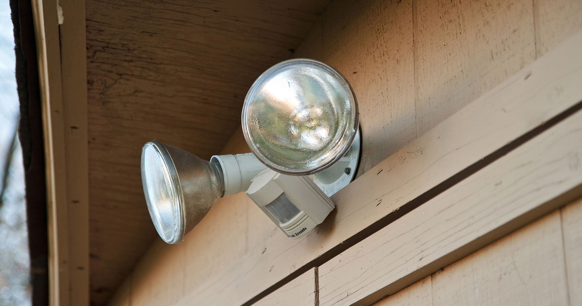 Two silver floodlights mounted on a beige building, with a motion sensor beneath them.