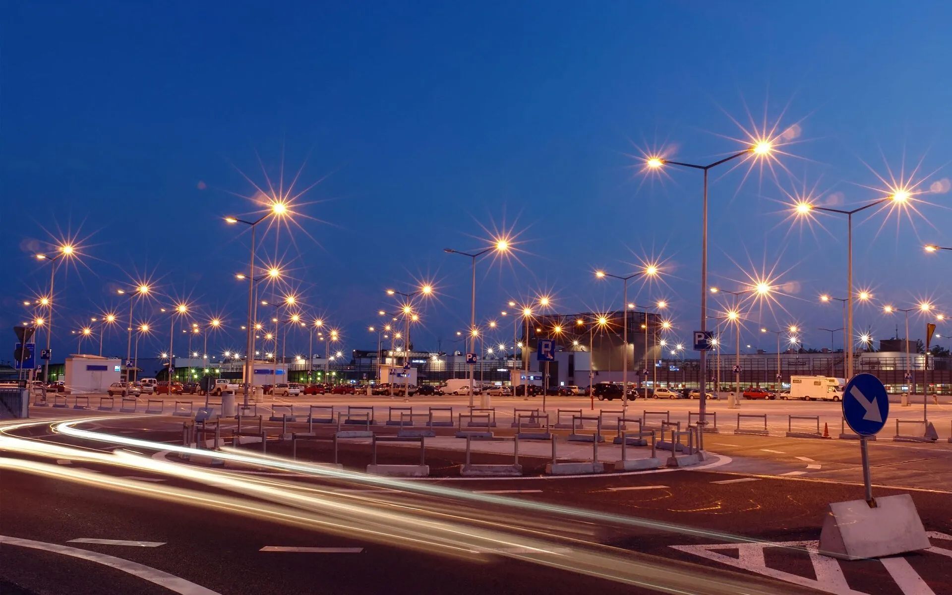 Night scene: busy airport parking lot with streetlights illuminating vehicles and buildings under a dark blue sky.