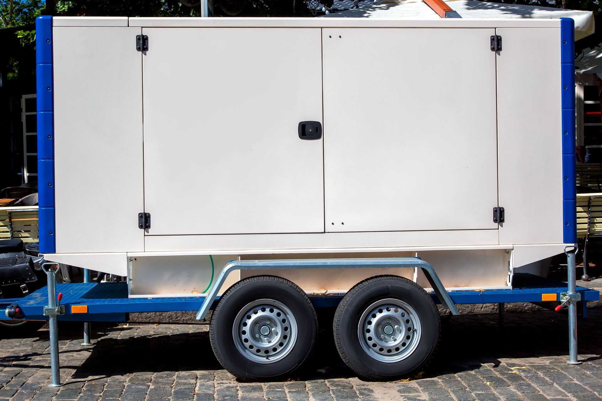 Generator enclosed in a white cabinet, mounted on a trailer with two wheels, parked on a paved surface.