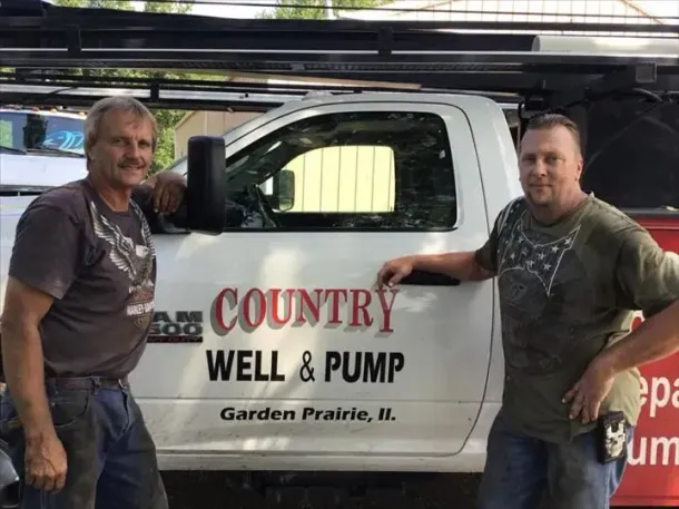 Two men standing by a white Country Well & Pump service truck in a garage.