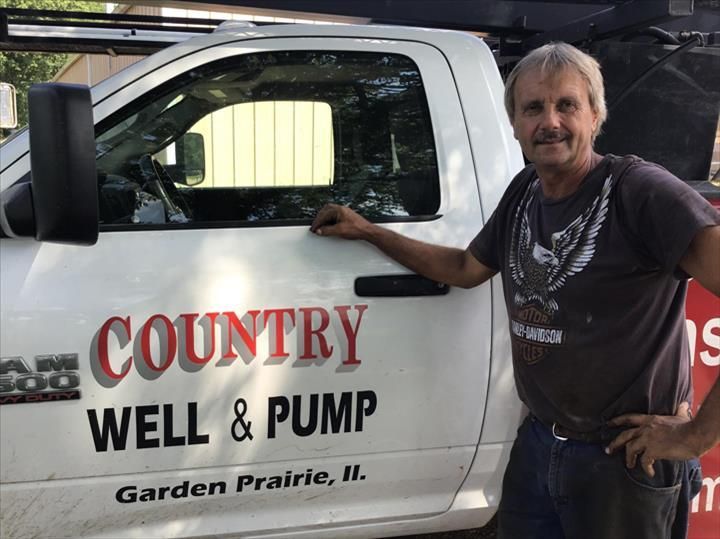 Man standing beside white Country Well & Pump truck with red lettering, garage setting