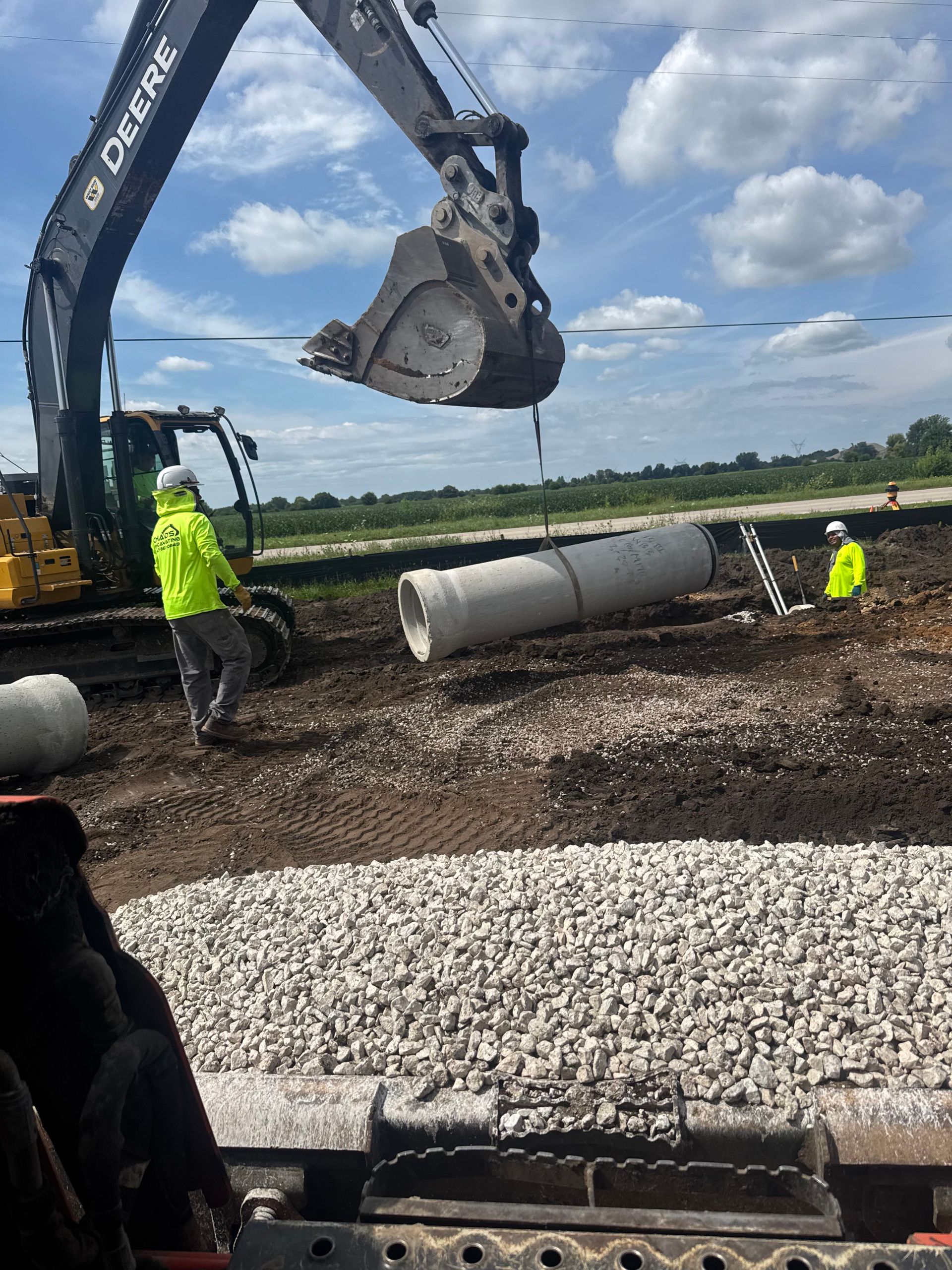 Excavator lifting a large concrete pipe at a construction site, with workers in neon safety gear nearby