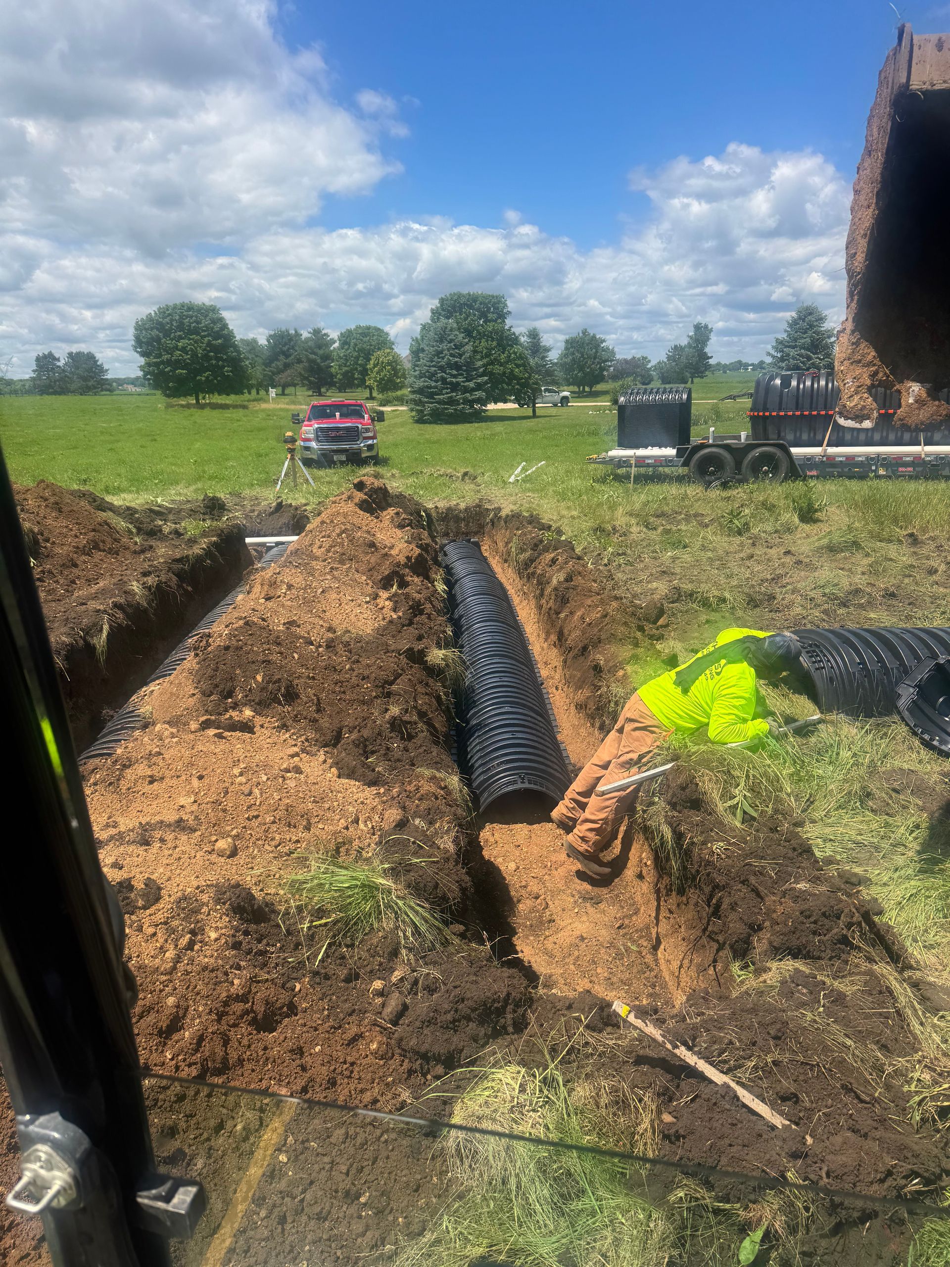 Excavation trench with black pipe and dirt piles in a grassy field under a cloudy sky, with machinery in the distance