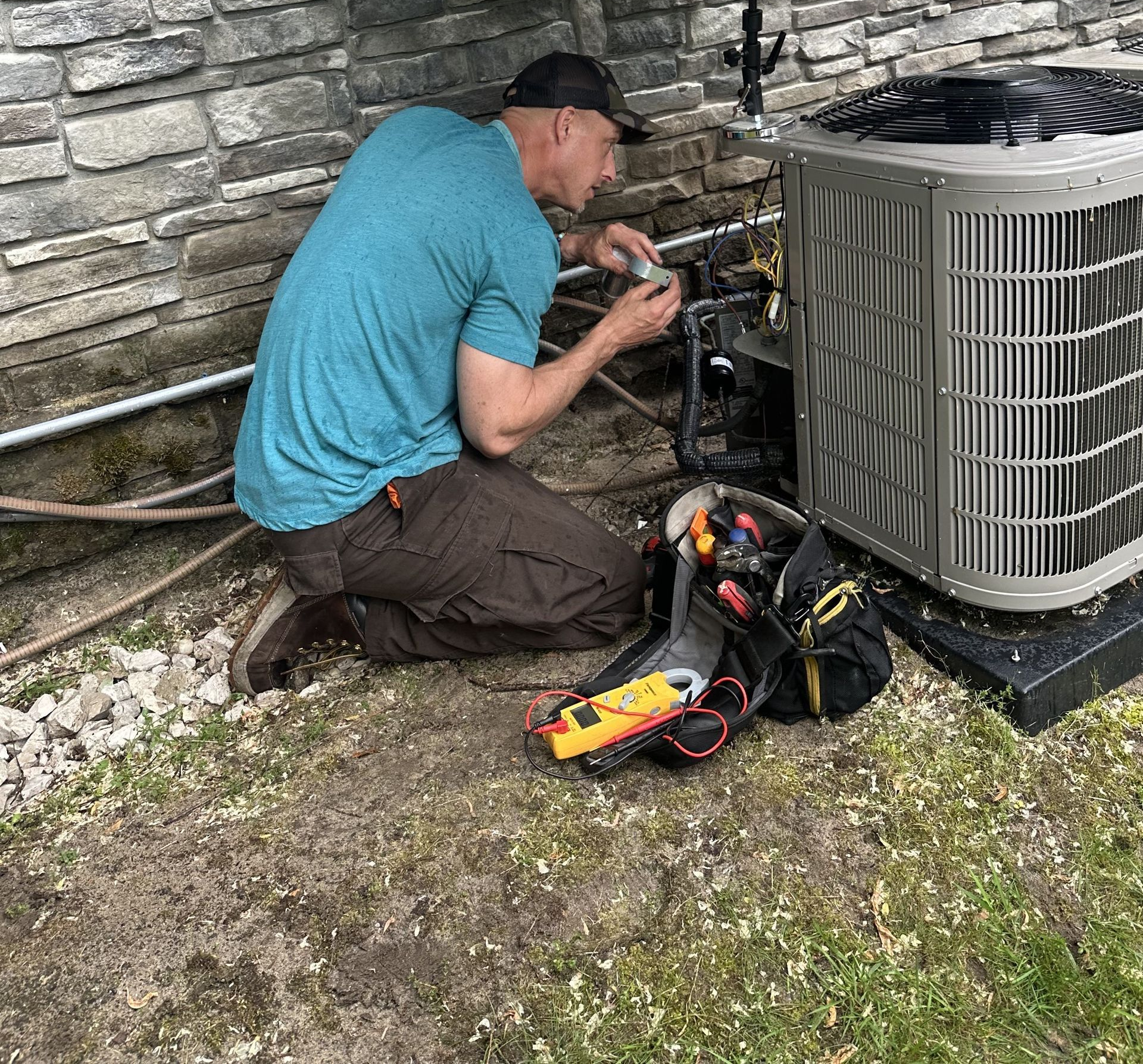 HVAC technician kneeling, working on AC unit next to stone wall, tool bag visible.
