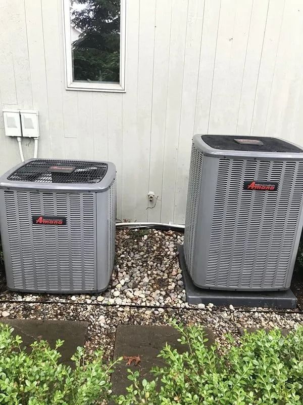 Two gray air conditioning units beside a light-colored building with a small window, set on gravel.