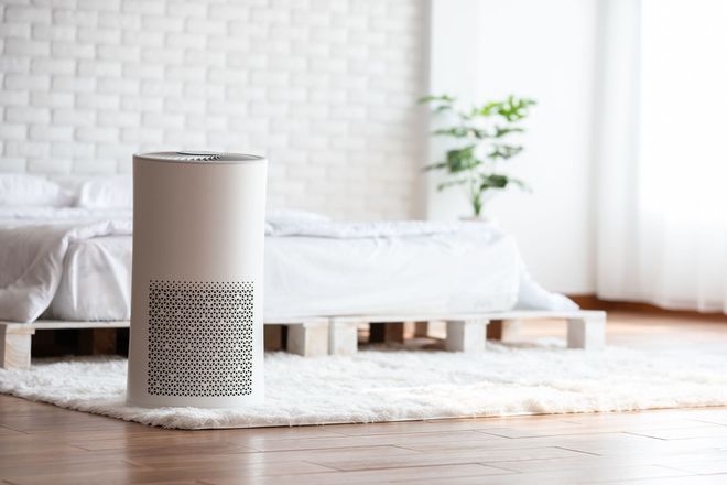 White air purifier in a bedroom, on a fluffy rug, near a bed and a plant.