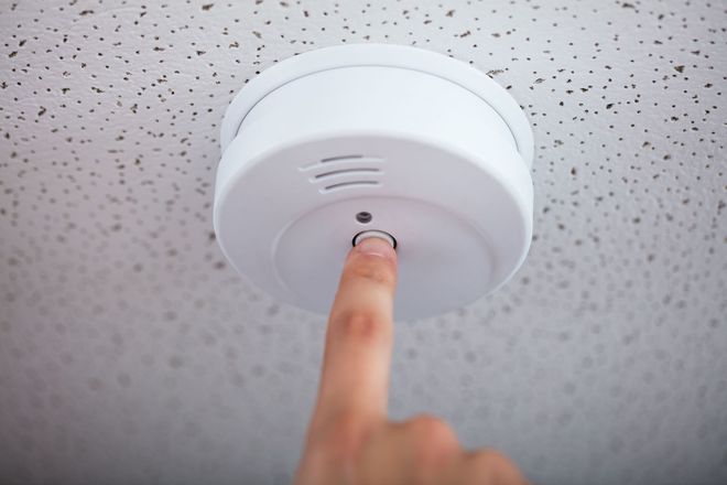 Finger pressing a button on a white smoke detector attached to a textured ceiling.