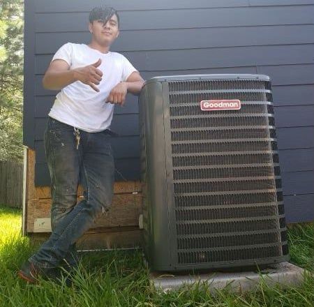 Person in a white shirt and jeans leaning on a Goodman air conditioning unit outdoors.