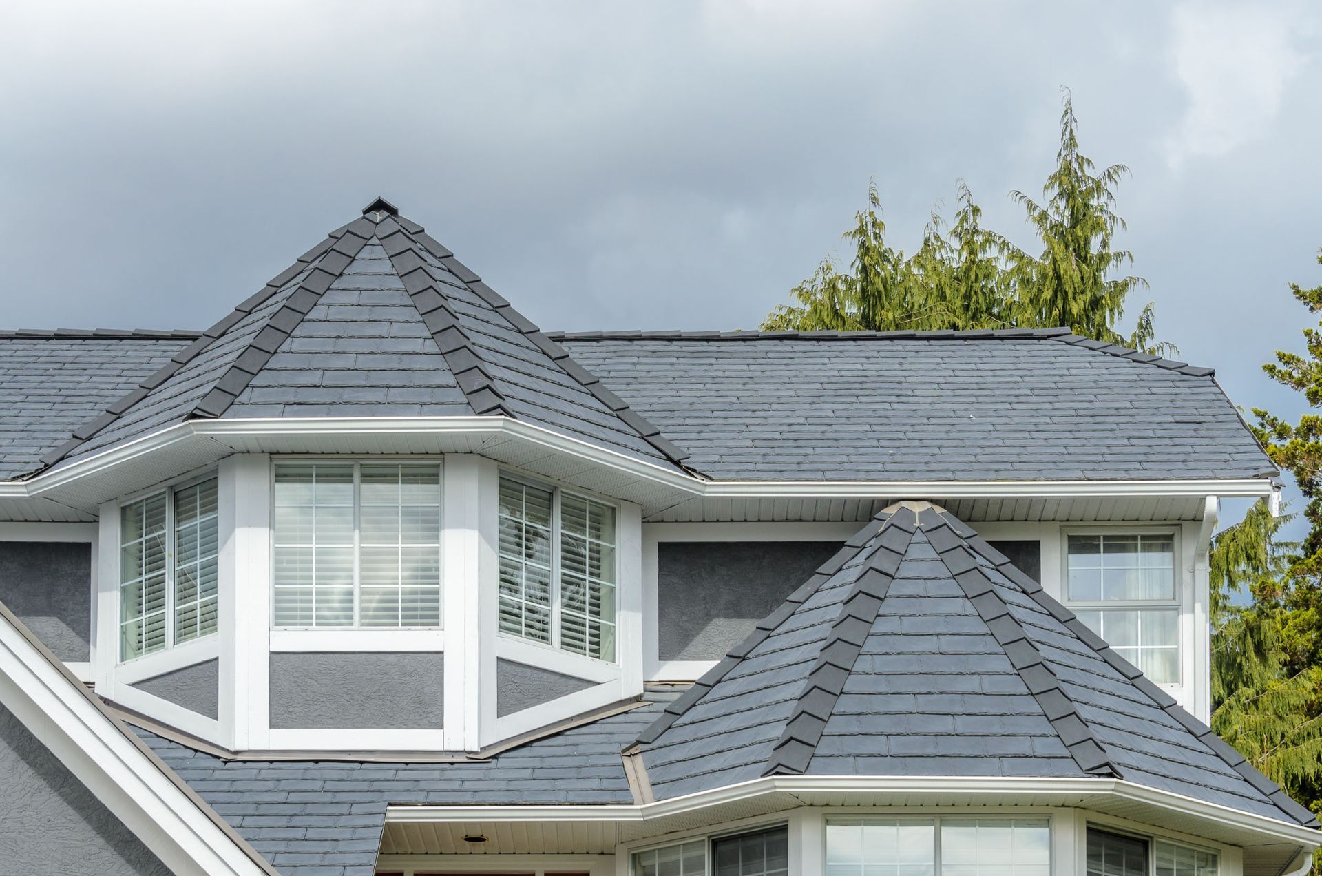 Gray house with dark gray roof, white trim, and bay windows against a cloudy sky.