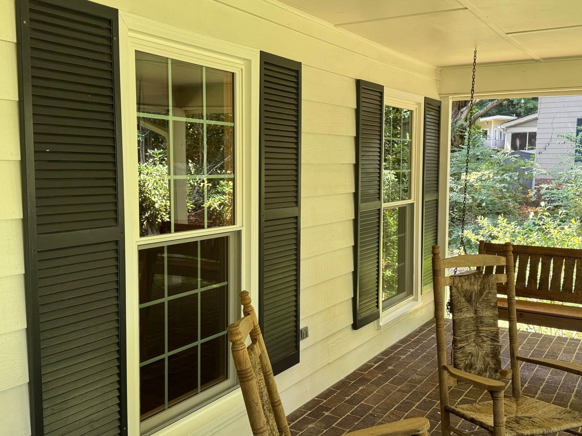 A porch with black shutters, a rocking chair, and a swing. Sunlight and greenery are visible.