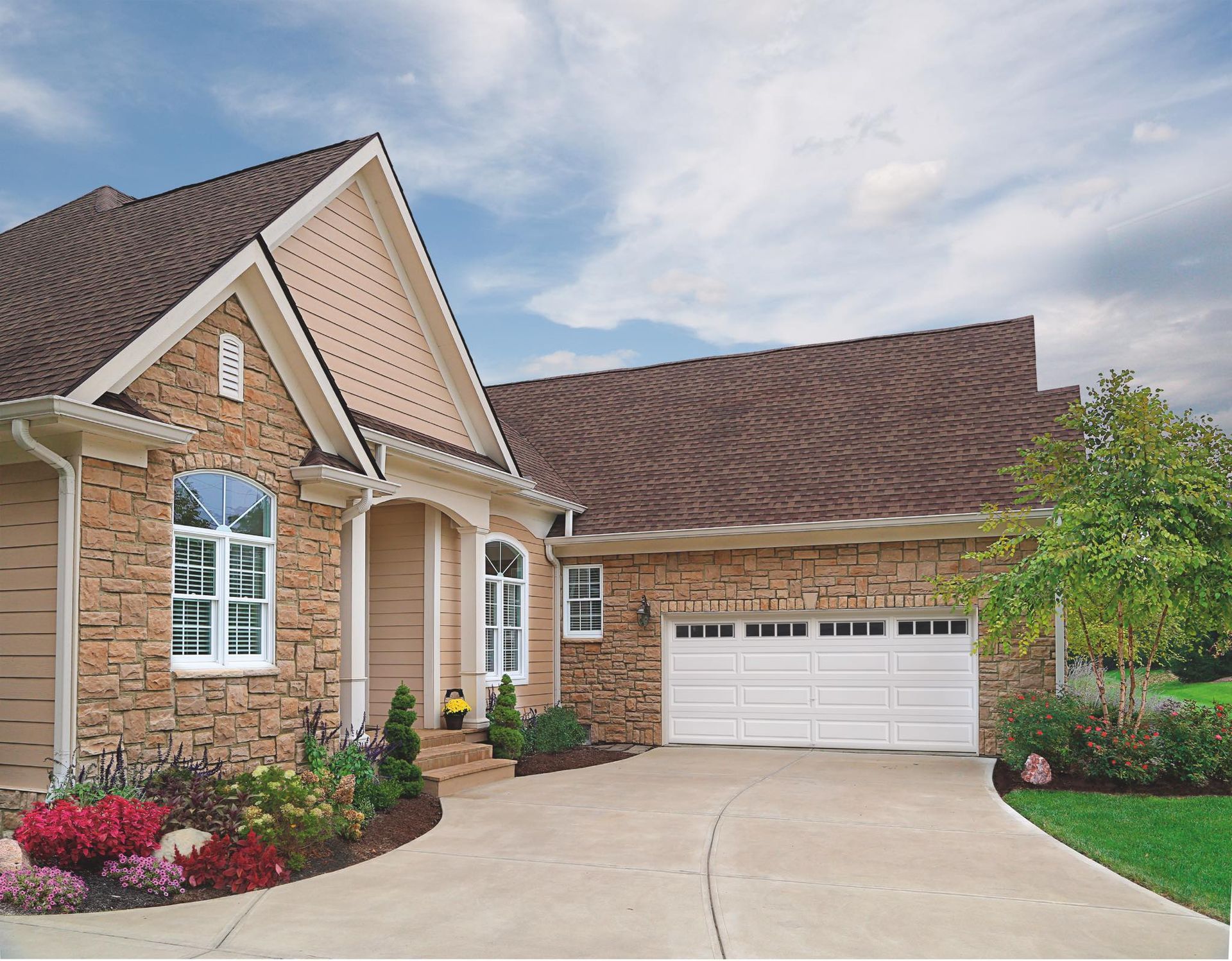 House with stone facade, driveway, garage, landscaping under cloudy sky.